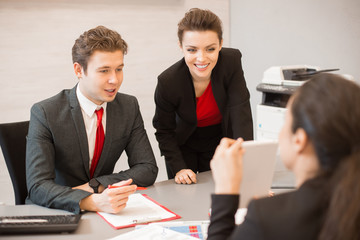 Portrait of three business people sitting at meeting table in modern office and discussing project plans, focus on business man and woman looking at statistic report and smiling happily