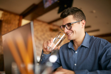 Smiling caucasian man with glass of water in hand works with laptop. Businessman in glasses drinks water for body hydration while working. Attractive designer quench thirst. Healthy lifestyle theme.