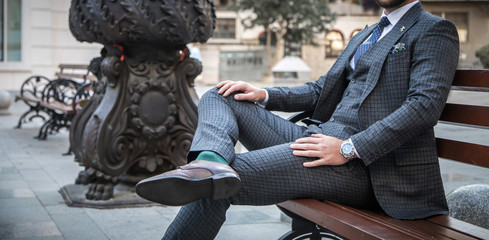 Suited man sitting on bench outdoors with his leg crossed and with his hands on his legs