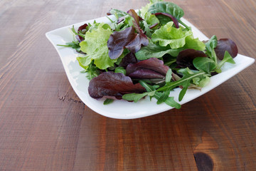 Fresh salad of mixed greens, mesclun, arugula, mache, lettuce, tender leaf vegetables in plate over wooden table