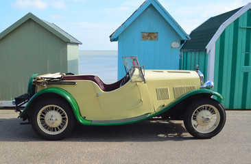Classic Cream & Green open top motor car parked on seafront promenade in front of beach huts.