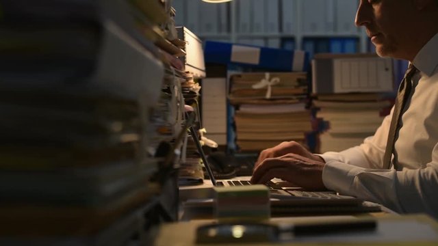 Businessman working with his laptop late at nigth, his desk is covered with paperwork, overtime work concept