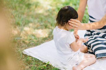 Rear crroped view of mother and her cute little daughter caring and playing outdoor on green grass. Cute mom and her child brush hair in the park. Portrait of happy family. Motherhood and childhood.