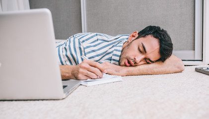 Young Caucasian asleep freelancer business man sleeping near to laptop with eyes closed while lying on the floor at home. Quick nap. People, lifestyle, technology, business and communication concept