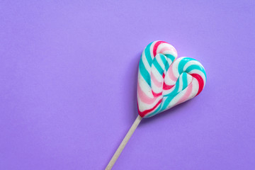View of a red and white and blue swirled heart shaped lollipop on purple background with copy space