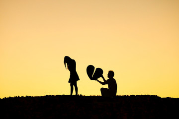 Man begging a woman for love by giving a big broken heart in sunset silhouette background...
