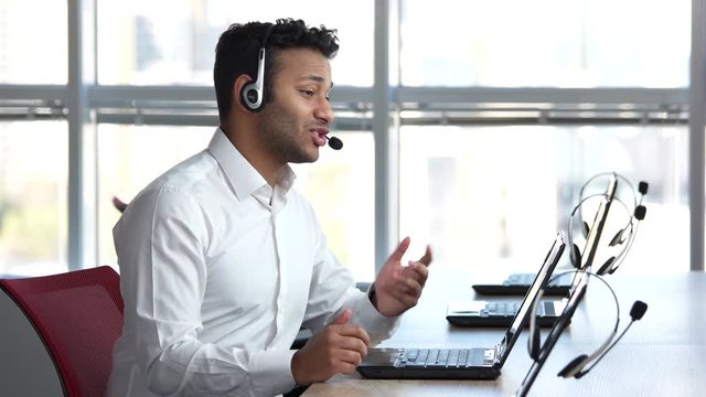 Indian man working in call center. Employeer in call center smiling and pleasantly answering client quesions, huge bright windows background.