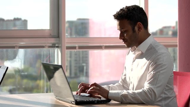 Indian smiling programmer working on laptop. Concept of indian man typing on laptop in frame office with city view out of window.