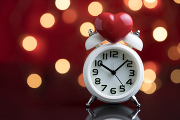 Obraz na płótnie Canvas Red heart shape on top of white alarm clock with party decoration light bokeh in the red background using as time for love or wake up after Valentines party