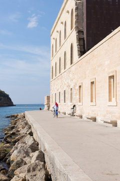 Woman Bicycling in Marseille. She is wearing a red hajib and has her back to the camera. She is on a sidewalk at the water's edge. 