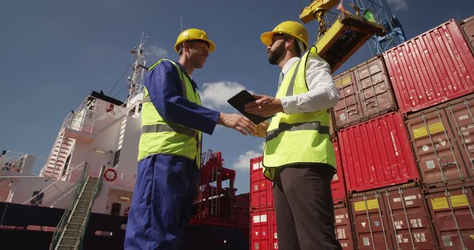 Dock workers shake hands and discuss shipping logistics in a shipyard. Shot on RED Epic.
