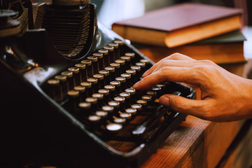 Human hands typing on vintage type writer machine and piles of books on wooden table - very selective focus.
