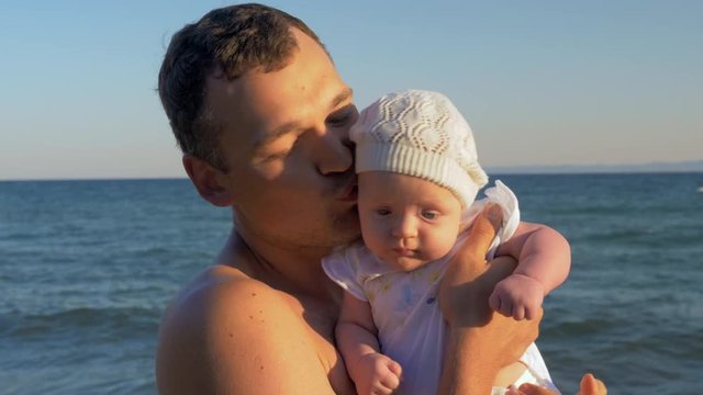 Young father with baby daughter in hands on sea background. Man kissing beloved child and looking to the camera. Happy fatherhood