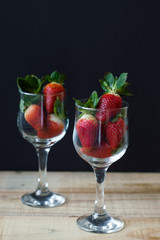 Two vine glasses with fresh strawberry with green leaves on wooden table, black background. Healthy dessert