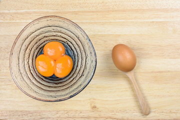 Egg yolk in glass bowl and raw egg in wood spoon on wooden table