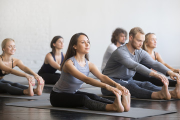 Group of young sporty people practicing yoga lesson with instructor, sitting in paschimottanasana...