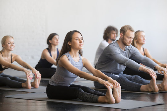 Group of young sporty people practicing yoga lesson with instructor, sitting in paschimottanasana exercise, Seated forward bend pose, working out, students training in fitness club, indoor, studio