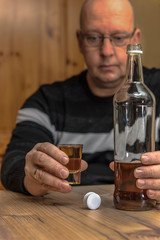 A man (45-55) is sitting at the table holding a glass of brandy in one hand and in the other hand she reaches for the brandy bottle. Concept: alcohol abuse or drinking