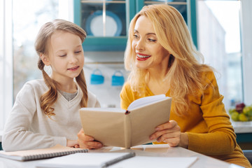 Funny reading. Pretty inspired blond slim mother smiling and looking at her daughter and while sitting at the table and her daughter reading