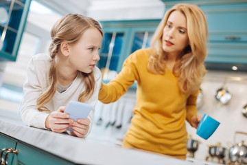Whats wrong. Pretty frustrated fair-haired girl holding a phone and her mom standing behind her and holding a cup of tea