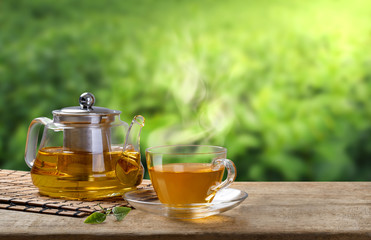 Warm cup and glass jugs or jars of tea and tea leaf and sack on the wooden table and the tea plantations background