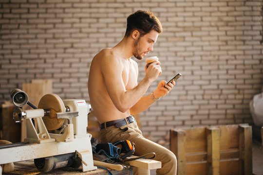 Carpenter Man using smart phone and holding cup of coffee in front of table with tools at workshop
