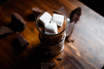 An underexposed horizontal image of hot chocolate in a glass in a metal glass-holder, decorated with marshmallows and pieces of dark chocolate on a wooden table. Selective focus.