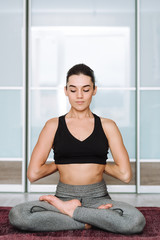 Beautiful brunette girl sitting in lotus pose. The model practices yoga in a bright room.