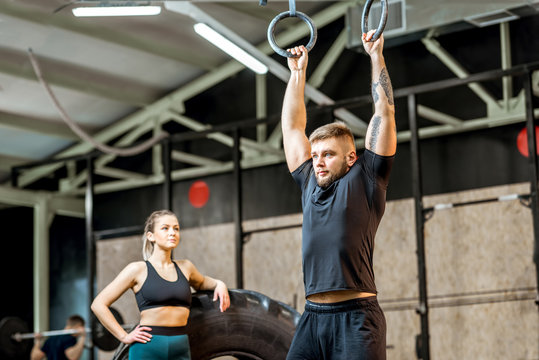 Handsome man in black sportswear pulling up on the gymnastic rings with woman on the background in the gym