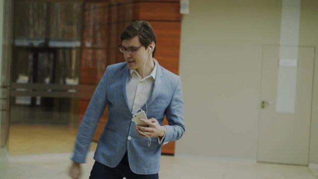 Happy businessman listening music with earphones dancing and walking in lobby of modern office