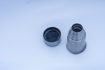 A thermos with tea stands on the snow. A metal mug in the winter in the forest.