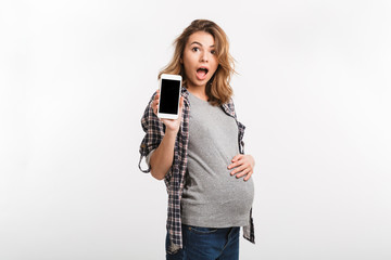 portrait of shocked pregnant woman showing smartphone with blank screen isolated on grey