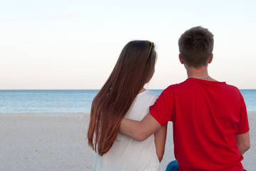 Fototapeta na wymiar romantic date girl with a guy sitting on the seashore, a guy hugs a girl by the shoulder, long hair for a woman and a red t-shirt for a man, on the beach and on a bench