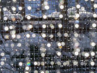 Background of weld mesh with melting snow creating droplets of sparkling water and round bokeh. Pretty glistening light effects. - obrazy, fototapety, plakaty