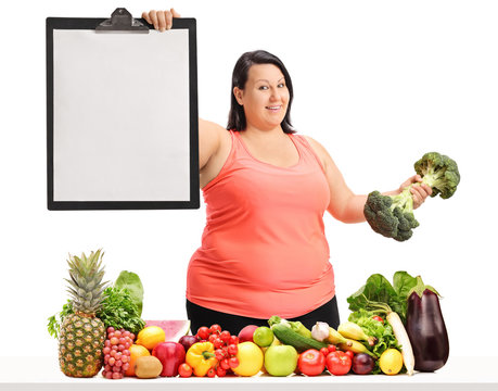 Overweight woman holding a blank clipboard and a broccoli dumbbell behind a table with fruit and vegetables