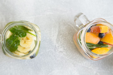 Two glass jars of detox infused water with sliced babanas, grapefruit, blueberry, rosemary and spinach, light concrete background, top view