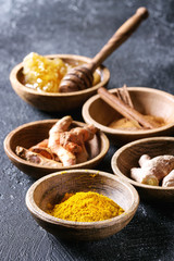 Ingredients for turmeric latte. Ground turmeric, curcuma root, cinnamon, ginger, honeycombs in wooden bowls over black texture background. Close up