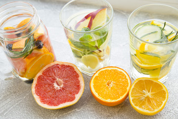 Three glasses of detox water with sliced fruit, vegetables, berries and herbs and fresh cut grapefruit and orange on a light stone background