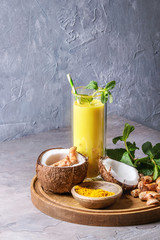 Cup of ayurvedic drink golden coconut milk turmeric iced latte with curcuma powder, crushed ice, mint and ingredients above on round wooden tray over grey kitchen table.