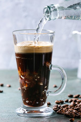 Fizzy iced Coffee espresso with pouring sparkling water from bottle and roasted beans around over grey green texture table. Modern drink.