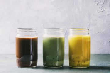 Fototapeta na wymiar Variety of iced colorful latte drinks. Iced coffee, turmeric and matcha latte cocktails in glass jars over grey green texture background. Copy space. Toned image