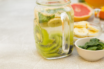 A glass jar of detox water with sliced banana, kiwi, and mint and bowl with ingridients on a light stone background