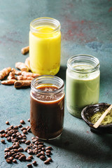Variety of iced colorful latte drinks. Iced coffee, turmeric and matcha latte cocktails in glass jars with ingredients above over grey green texture background. Copy space