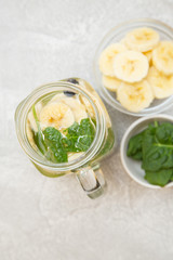 A glass jar of detox water with sliced banana, kiwi, and mint and bowl with ingridients on a light stone background, top view