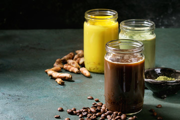 Variety of iced colorful latte drinks. Iced coffee, turmeric and matcha latte cocktails in glass jars with ingredients above over grey green texture background. Copy space