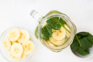 A glass jar of detox water with sliced banana, a bowl of spinach and a plate with banana slices on white, top view