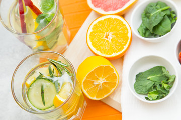 Two glasses of detox water with sliced cucumbers, lemon and apple, sninach and mint in ceramic bowls and cut lemon, orange and grapefruit on a table with cutting boards and white napkin