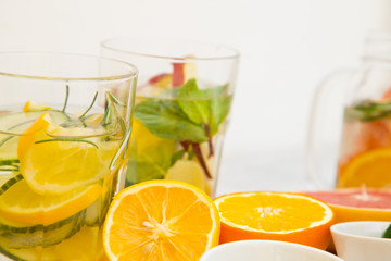 A close up of glasses of detox water with sliced lemon, cucumber and rosemary and cut oranges on the table, white background