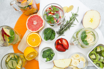 Glasses of detox water with sliced fruit, vegetables, berries and herbs, set of ingredients: banana, strawberry, orange, grapefruit, mint, spinach and rosemary on cutting boards and napkin, top view