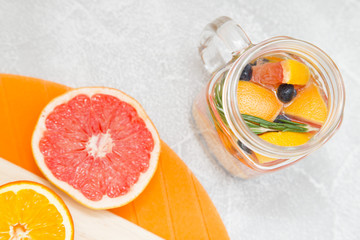 Detox infused water with slices of grapefruit, orange, blueberry and rosemary, halves of orange and grapefruit, light stone and cutting boards as a background